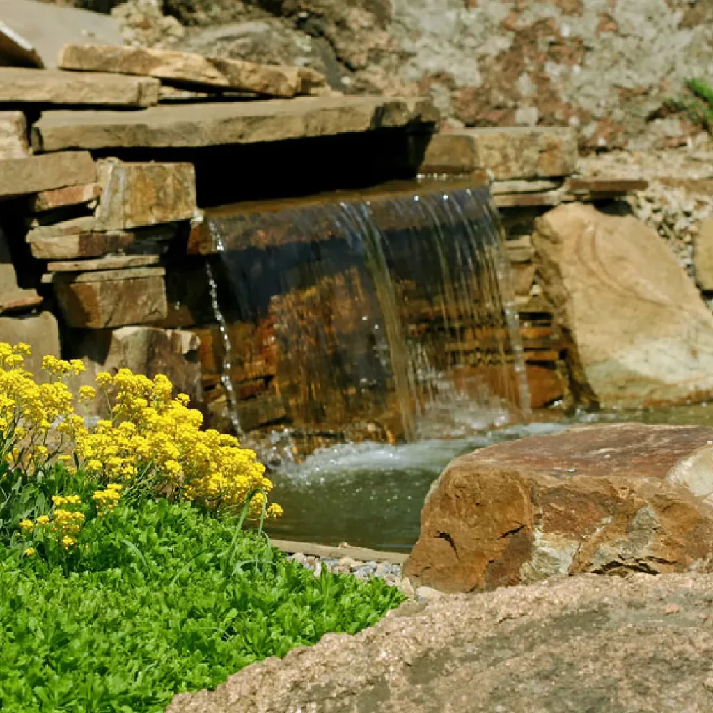 Backyard waterfall cascading over stacked stone ledge into a pond with yellow wildflowers and natural boulders.