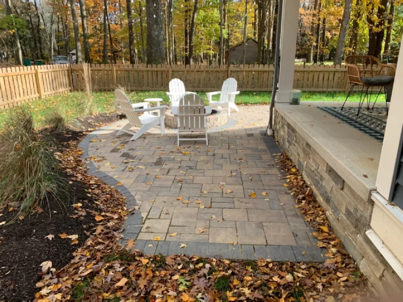 Paver patio in a fenced in wooded backyard with a custom stone firepit and patio chairs.