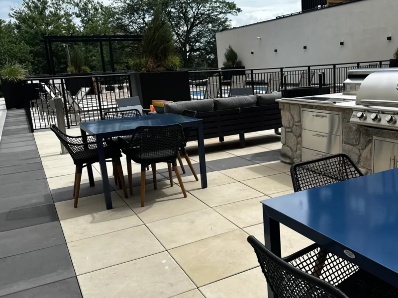 An outdoor lounge area with custom paver patio around a sunlit pool with an outdoor kitchen cooking area.