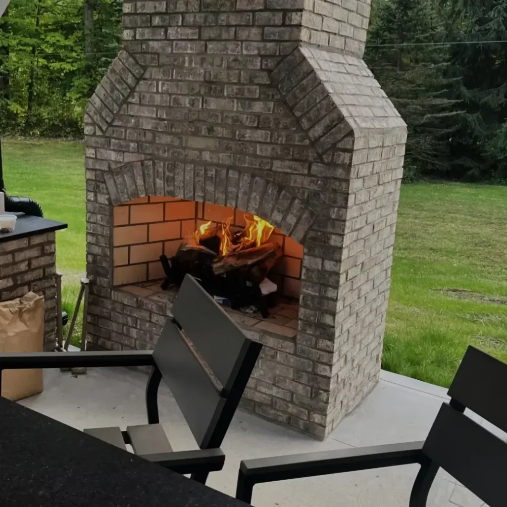 Custom brick outdoor fireplace with arched masonry firebox and open wood fire burning on a backyard patio.