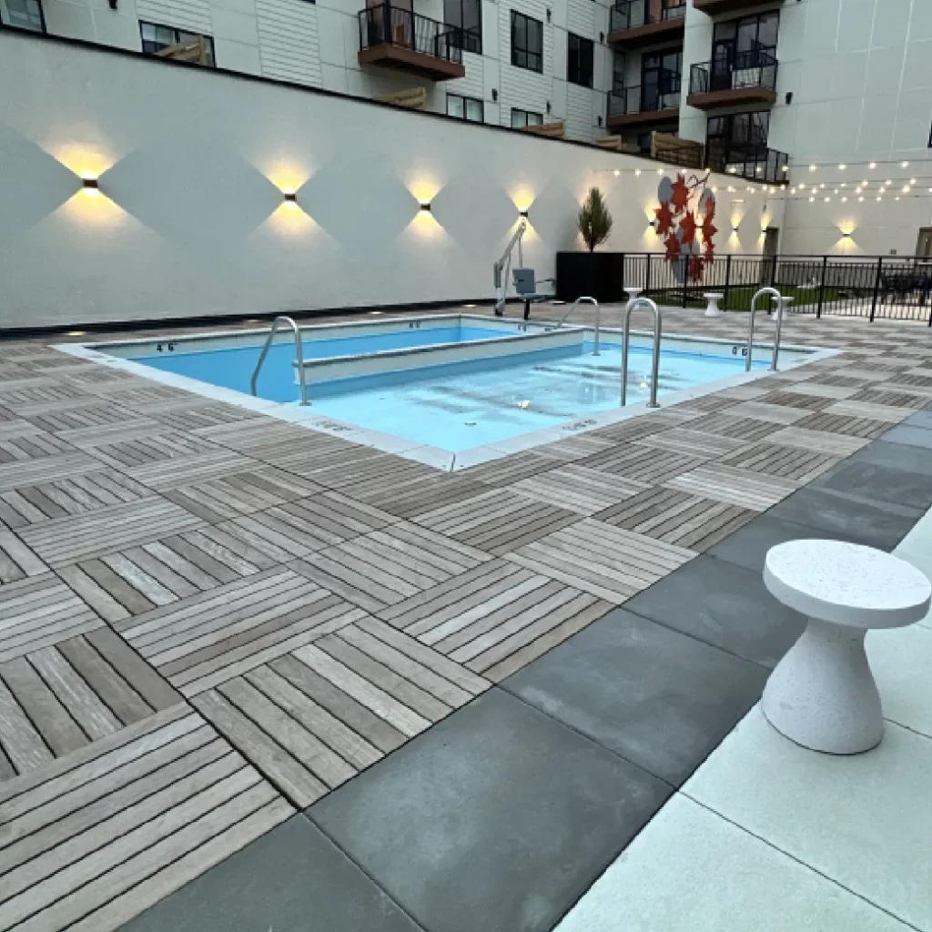 Custom pool deck lighting with wall-mounted uplights, string lights, and wood tile deck surrounding a residential pool.