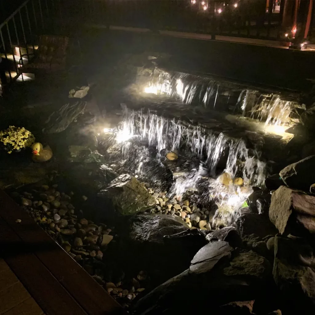 Outdoor lighting highlights a backyard waterfall and pond feature cascading over natural stone at night.