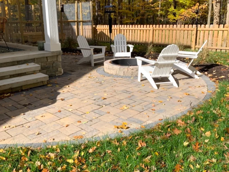 Paver patio installation with fire pit and seating area in fenced, wooded yard in autumn.
