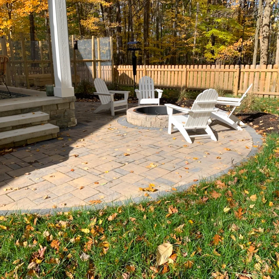 Paver patio installation with fire pit and seating area in fenced, wooded yard in autumn.