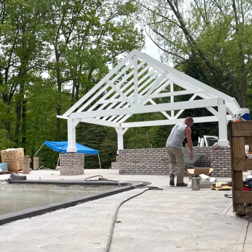 Pergola construction project in progress over a pool deck with brick knee walls and craftsman at work.