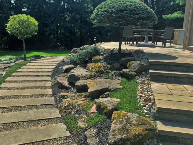 Stone patio installation with large slab walkway, stone steps, natural boulder garden, and manicured trees.