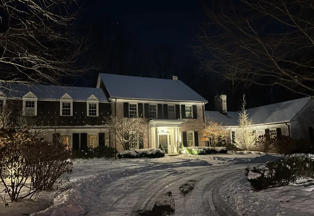 A large colonial home in Chagrin Falls shows why the color brown is still beautiful in winter, with different plants and decorative trees in a front yard, lit at night by professionally installed outdoor lighting