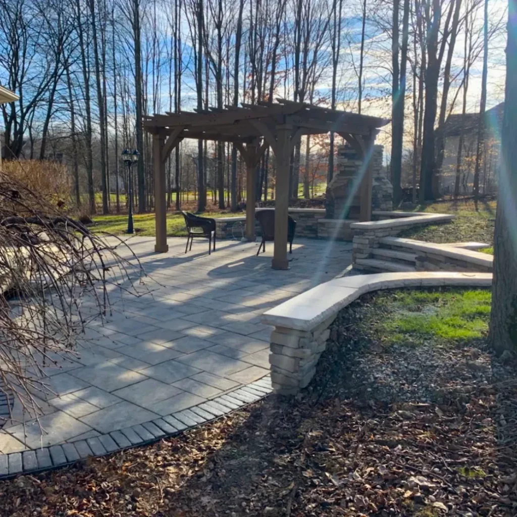 Wood pergola construction with paver patio and stone retaining wall on a wooded residential property.